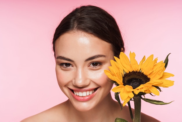 Schönheitsporträt einer schönen lächelnden jungen topless Brunettefrau, die lokalisiert auf Rosa steht und Sonnenblume hält