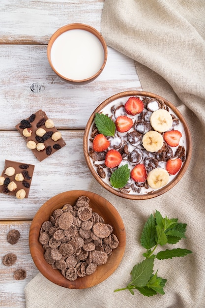 Schokoladen-Cornflakes mit Milch und Erdbeere in der Holzschale auf weißer Holzoberfläche und Leinentextil