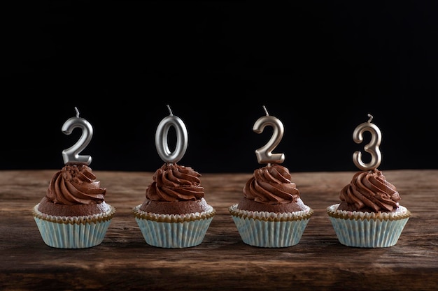 Schokoladen-Cupcakes mit Buttercreme-Zuckerguss mit silbernen 2023-Kerzen auf Holztisch auf schwarzem Hintergrund