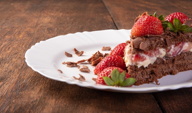 Schokoladenkuchen, ein leckeres Stück Schokoladenkuchen mit Erdbeeren auf einem weißen Teller auf rustikalem Holz, abstrakter Hintergrund, selektiver Fokus.