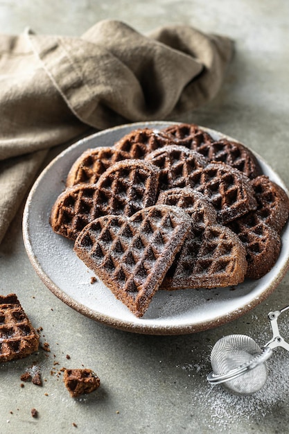 Schokoladenwaffeln in Form eines mit Puderzucker bestreuten Herzens in Keramikplatte auf strukturiertem Hintergrund mit einer Leinenserviette im Vordergrund, Stillleben zum Valentinstag