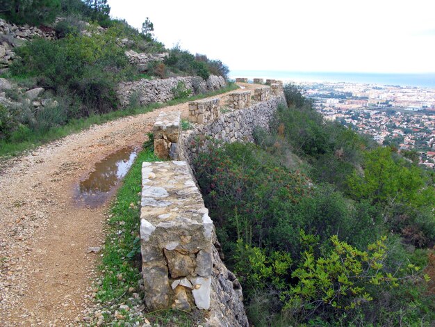 Schotterlehmstraße bergauf, altes Mauerwerk auf der Seite Panorama der Stadt unten, Berg Montgo Denia Spanien