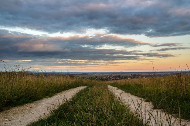 Schotterstraße zwischen dunklen Feldern bei Sonnenuntergang mit dramatischer Wolkenlandschaft.