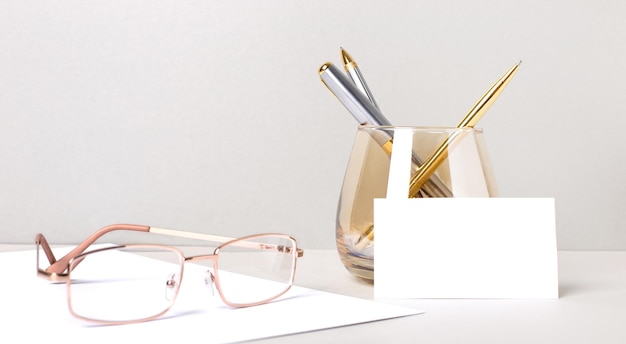 Schreibtisch mit goldener Brille, Stiften in einem Glas und weißer leerer Karte mit Platz zum Einfügen von Text. Geschäftsvorlage. Heimbüro. Arbeitsplatz Nahaufnahme.