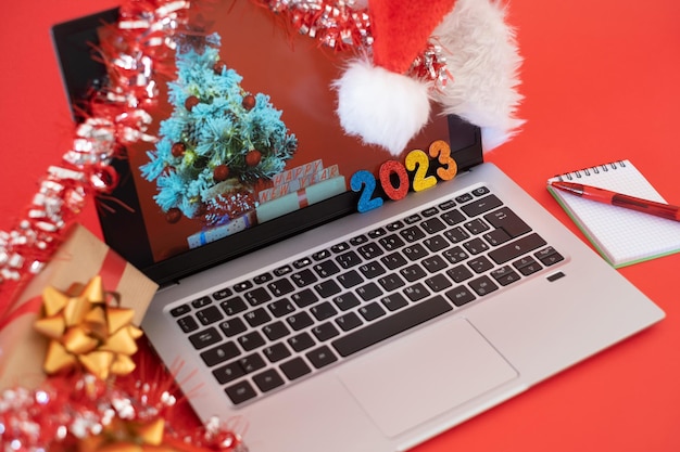 Schreibtisch mit Laptop zur Weihnachtszeit Botschaft des guten Rutsch ins neue Jahr 2023 roter Hintergrund mit Weihnachtsgeschenken und Weihnachtsmütze