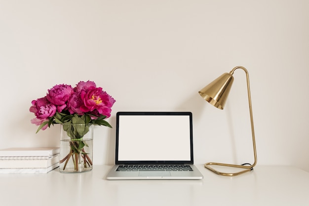 Schreibtischarbeitsplatz mit leerem Kopierraum Laptop-Bildschirm. Modernes stilvolles Hauptinnendesign mit Blumenstrauß der rosa Pfingstrosen