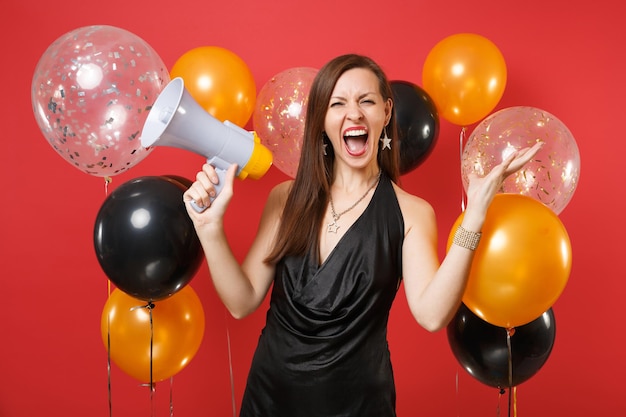 Schreiende junge Frau im schwarzen Kleid, die feiert, Megaphon hält und Hände auf Luftballons mit rotem Hintergrund ausbreitet. Internationaler Frauentag, Happy New Year Geburtstagsmodell Urlaub Party Konzept.