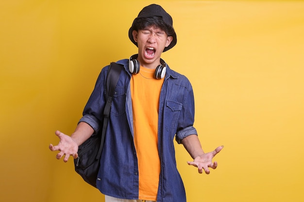 Schreiender junger asiatischer Mann mit Rucksack und Kopfhörern, der mit aggressivem Ausdruck die Hände hebt