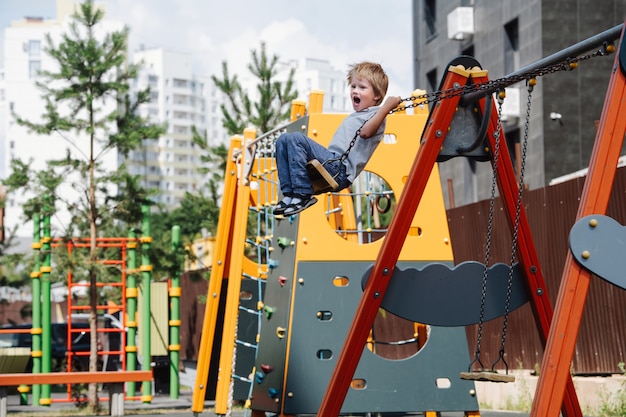 Schreiender kleiner Junge, der auf Schaukeln auf einem Kinderspielplatz reitet. Er fliegt hoch und biegt Kettengriffe.