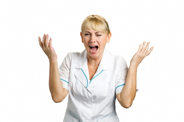 Schreiender medizinischer Arbeiter auf Weiß. Frustrierte und schockierte reife Krankenschwester mit erhobenen Händen und offenem Mund, der auf Weiß steht.