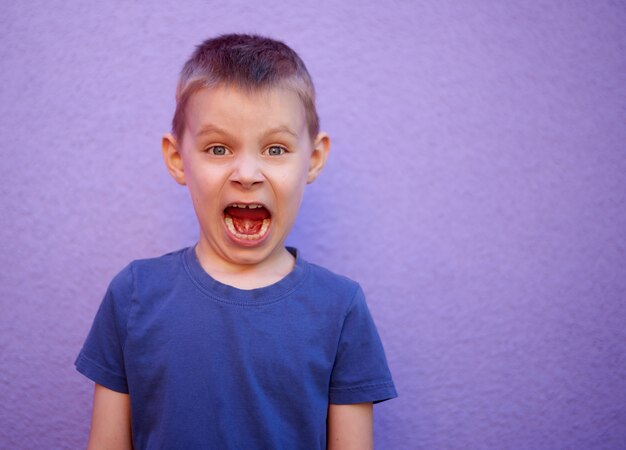 Schreiender sechsjähriger Junge in einem blauen T-Shirt auf violettem Hintergrund