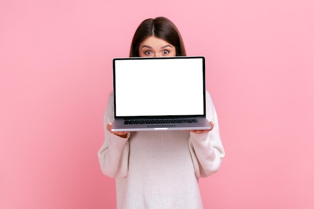 Schüchterne brünette Frau, die ein Notizbuch in Händen hält, das die Hälfte des Gesichts bedeckt, mit einem Laptop, der späht und einen weißen, lässigen Pullover trägt. Studioaufnahme im Innenbereich isoliert auf rosa Hintergrund