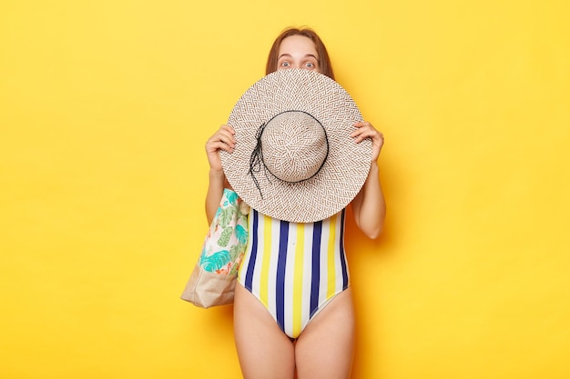 Foto schüchterne frau im gestreiften badeanzug isoliert auf gelbem hintergrund versteckt ihr gesicht hinter ihrem strohhut und steht mit einer tasche im sommerurlaub