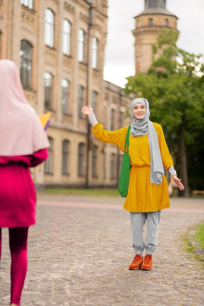 Schüler winken. Muslimischer Student mit grüner Handtasche winkt, während er einen Freund in der Nähe der Universität trifft