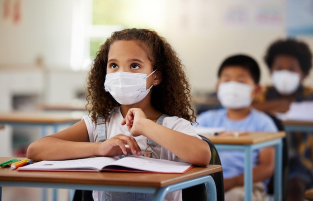 Schülerin mit Covid-Lernen im Unterricht, die eine Maske trägt, um sich vor Viren zu schützen, und auf die Bildung im Klassenzimmer konzentriert ist Kleines Mädchen, das am Schreibtisch sitzt und während einer Pandemie lernt und zuhört