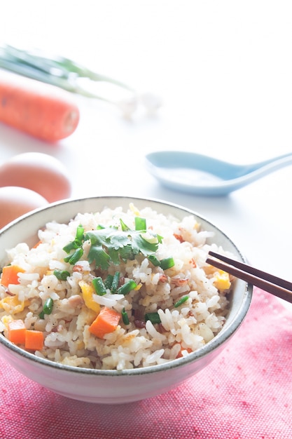 Schüssel gebratener Reis, Karotten und Ei auf rosa Stoff, Löffel, Stäbchen und Zutaten für Hintergrund