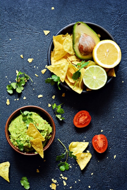 Schüssel Guacamole Dip mit Mais Nachos (Chips) und Zutaten auf dunklem Hintergrund, selektiver Fokus.