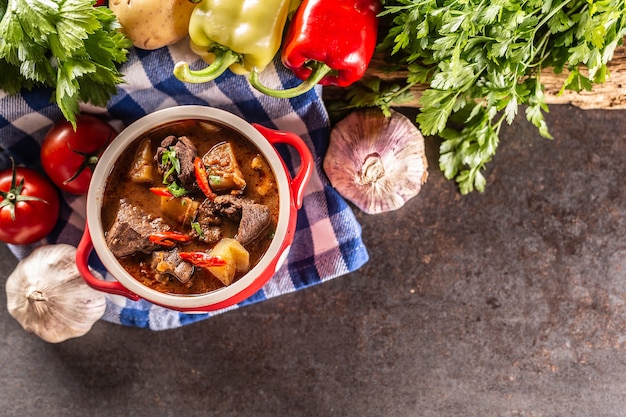 Schüssel Gulaschsuppe mit Fleisch, Kartoffeln und Chilis neben rohen Zutaten für den Eintopf.