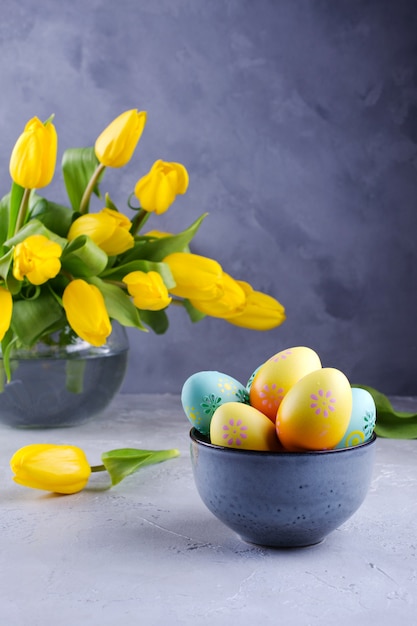 Schüssel mit bunten Ostereiern; Frühlings-Osterndekoration auf grauem Tisch mit Strauß gelber Tulpenblüten in Glasvase; Ostern Innendekoration