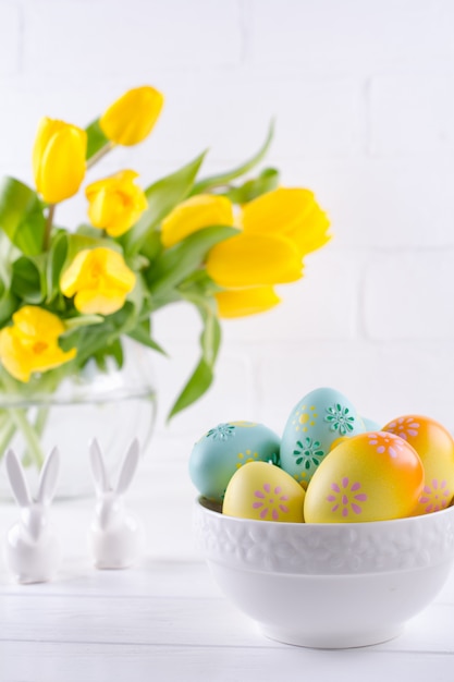 Schüssel mit bunten Ostereiern, Frühlings-Osterndekoration auf weißem Holztisch mit Strauß gelber Tulpenblumen in Glasvase auf weißem Hintergrund. Ostern Innendekoration