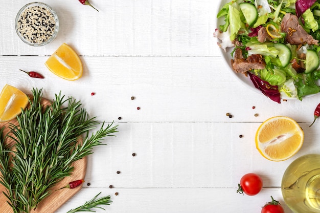 Schüssel mit frischem Salat, Rosmarin, Zitrone, Gewürzen und Öl auf weißem Holztisch. Lebensmittelhintergrund.