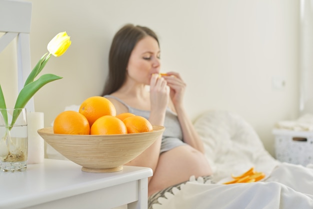 Schüssel mit Orangen im Fokus, schwangere junge Frau, die Orangenscheiben isst, die im Bett sitzen