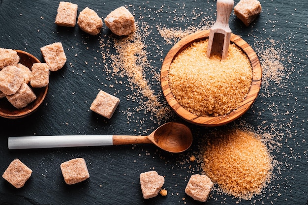 Schüssel und Schaufel mit Sand und Klumpen Brauner Zucker auf Holzhintergrund Brauner Zuckerwürfel Machen Sie ungesunde Ernährung, Fettleibigkeit, Diabetes, Zahnpflege und vieles mehr