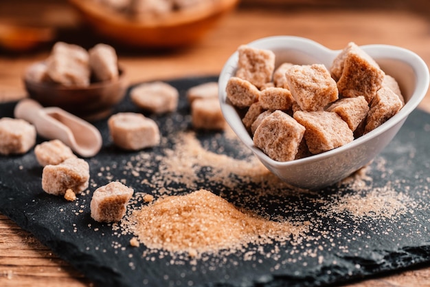 Schüssel und Schaufel mit Sand und Klumpen Brauner Zucker auf Holzhintergrund Brauner Zuckerwürfel Machen Sie ungesunde Ernährung, Fettleibigkeit, Diabetes, Zahnpflege und vieles mehr