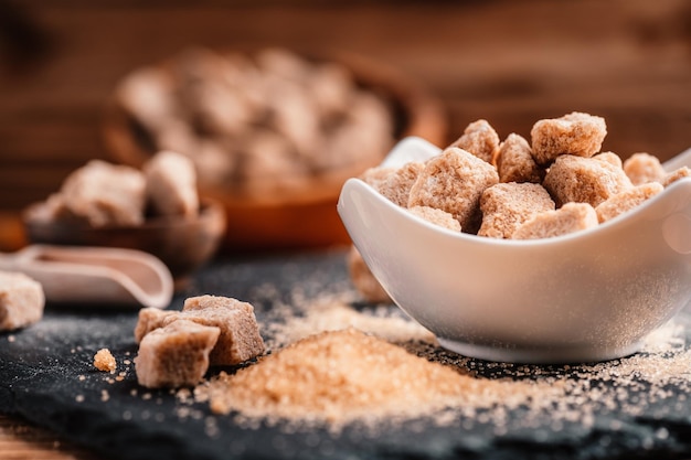 Schüssel und Schaufel mit Sand und Klumpen Brauner Zucker auf Holzhintergrund Brauner Zuckerwürfel Machen Sie ungesunde Ernährung, Fettleibigkeit, Diabetes, Zahnpflege und vieles mehr