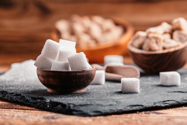 Schüssel und Schaufel mit weißem Sand und Klumpen weißer Zucker auf Holzhintergrund Zuckerwürfel Machen Sie ungesunde Ernährung, Fettleibigkeit, Diabetes, Zahnpflege und vieles mehr