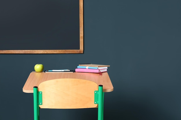 Schulbank aus Holz mit Schreibwaren und Apfel in der Nähe der Tafel an der grauen Wand