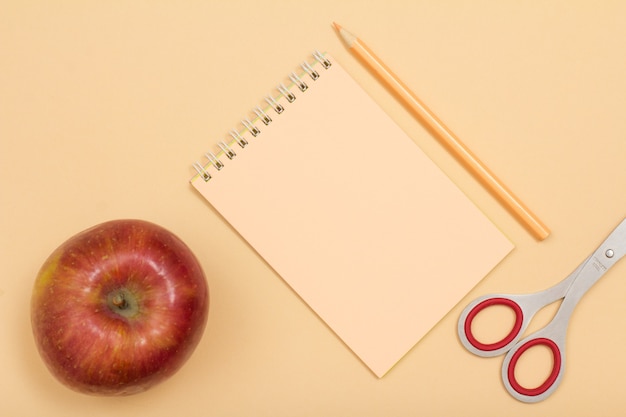 Schulbedarf. Apple, Notizbuch, Farbstift und Schere auf beigem Hintergrund. Ansicht von oben. Zurück zum Schulkonzept. Pastellfarben