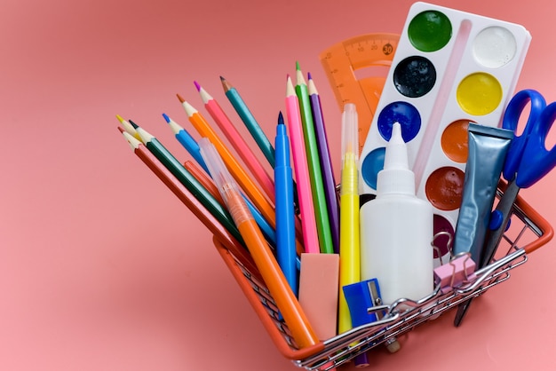 Schulbedarf in einem Einkaufskorb auf rosa Hintergrund. Vorbereitung auf die Schule, Kauf von Büromaterial. Zurück zur Schule.