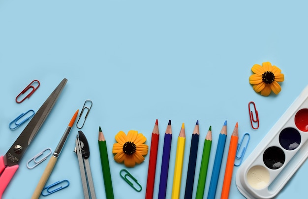 Schule stellte mit Bleistiften, Farben, Büroklammern, Bürsten, Scheren, Herbstblumen auf einem blauen Hintergrund ein