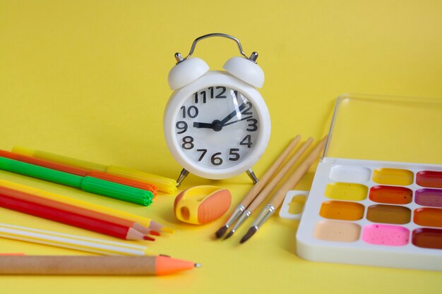 Schulhausaufgaben, Wecker, Bleistifte, Marker, Notizbuch, Kugelschreiber, Lineal, Spitzer, Aquarelle und Pinsel auf einem gelben Tisch