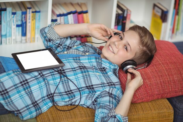 Schuljunge, der auf Couch entspannt, während Musik auf digitalem Tablett in Bibliothek hört