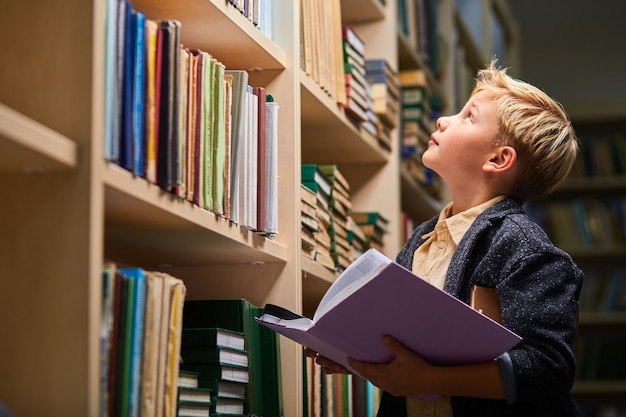 Schuljunge, der bücherregale in der universitätsbibliothek hochschaut, kinderjunge, der buch in händen hält