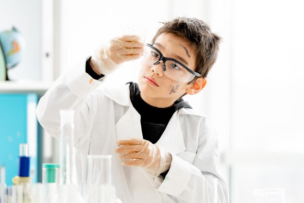 Schuljunge mit Schutzbrille, der während des Chemieexperiments im naturwissenschaftlichen Grundunterricht Flüssigkeit im Glas anschaut Kluger Schüler im Labor während des Tests