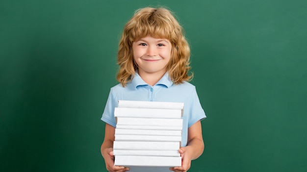 Schulkind in der Klasse Glückliches Kind gegen grüne Tafel Bildungs- und Kreativitätskonzept Kind, das Stapel Bücher mit Mörtelbrett auf Tafel hält