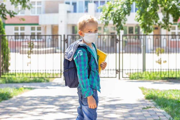 Schulkinder in einer Schutzmaske mit einem Rucksack und einem Lehrbuch in den Händen. in einem T-Shirt und einem karierten Shirt