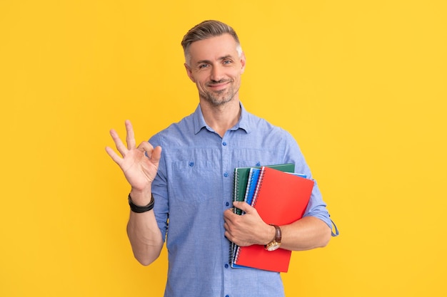 Schullehrer oder Universitätstutor glücklicher Mann halten Ordner mit ok Geste reifer Mann auf gelbem Hintergrund mit Projekttyp mit Dokumenten in der Zwischenablage, der einen Vertrag unterzeichnet