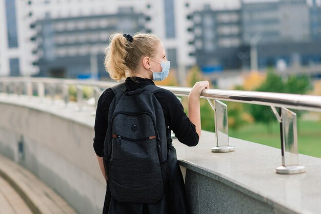 Schulmädchen in der medizinischen Schutzmaske bei Sonnenuntergang. Moderner Schüler mit Rucksack während der COVID-19-Quarantäne.