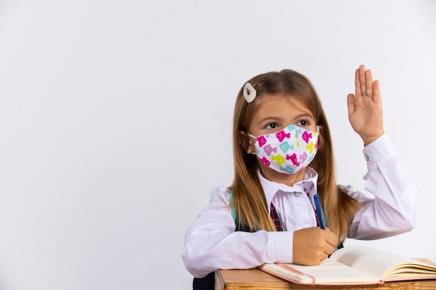 Schulmädchen in medizinischer Maske hob im Unterricht die Hände und trainierte Bücher auf dem Tisch. Schulkonzept