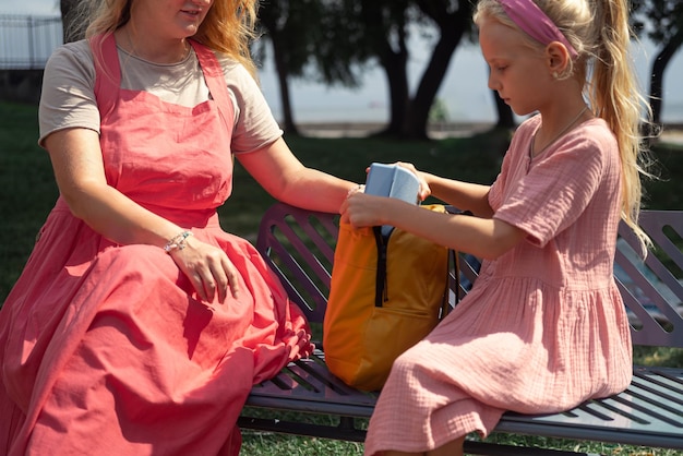 Schulmädchen in rosa Kleid steckt Lunchbox in Rucksack und sitzt mit junger Mutter auf Parkbank