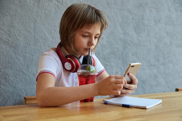 Schulmädchen mit Kopfhörern und roter eisiger Limonade nutzt Smartphone surft im Internet