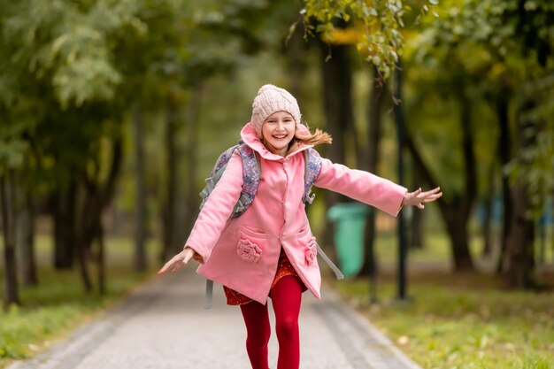 Schulmädchen mit Rucksack, das im Herbstpark läuft und lächelndes jugendliches Kind hat, das Spaß im Freien hat