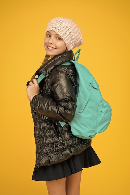 Schulmädchen-Mode-Outfit Kind mit Rucksack Modegeschäft Modekonzept Mädchen tragen Strickmütze und Jacke gelben Hintergrund Entzückende Schülerin Herbst Herbst Winter Kaufen Sie Kleidung für die Schulsaison