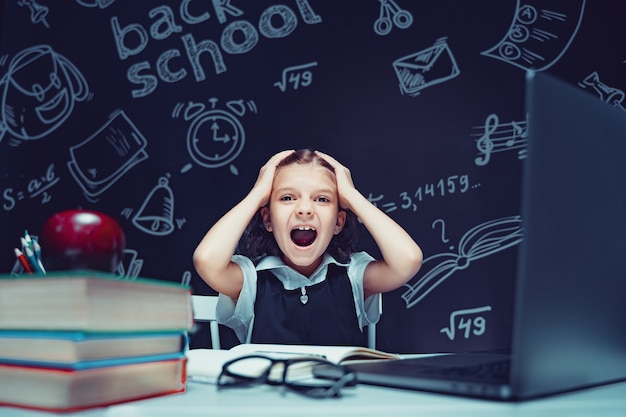 Schulmädchen sitzt am Tisch mit Laptop-Büchern und einer Tafel mit Schulformeln im Hintergrund...