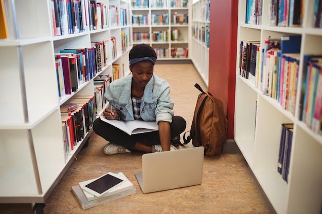 Schulmädchen sitzt auf dem Boden und macht Hausaufgaben in der Bibliothek