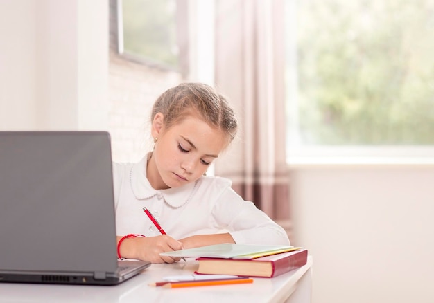 Schulmädchen sitzt mit einem Laptop an einem Tisch und schreibt in ein Notizbuch